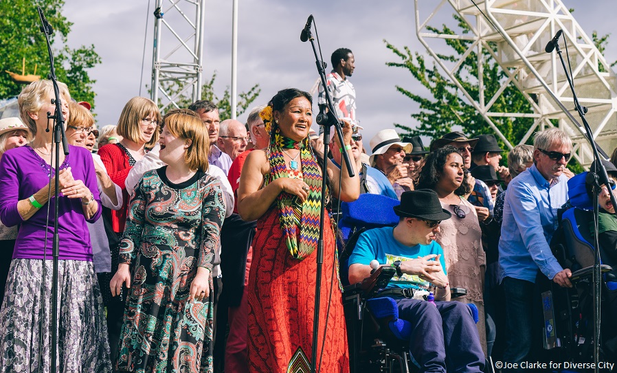 A group of people outdoors in front of microphones, taking part in an artistic event, including people of colour and a wheelchair-user