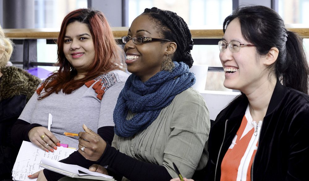 Three women of different ages and ethnicities sit smiling in a row with notebooks, learning