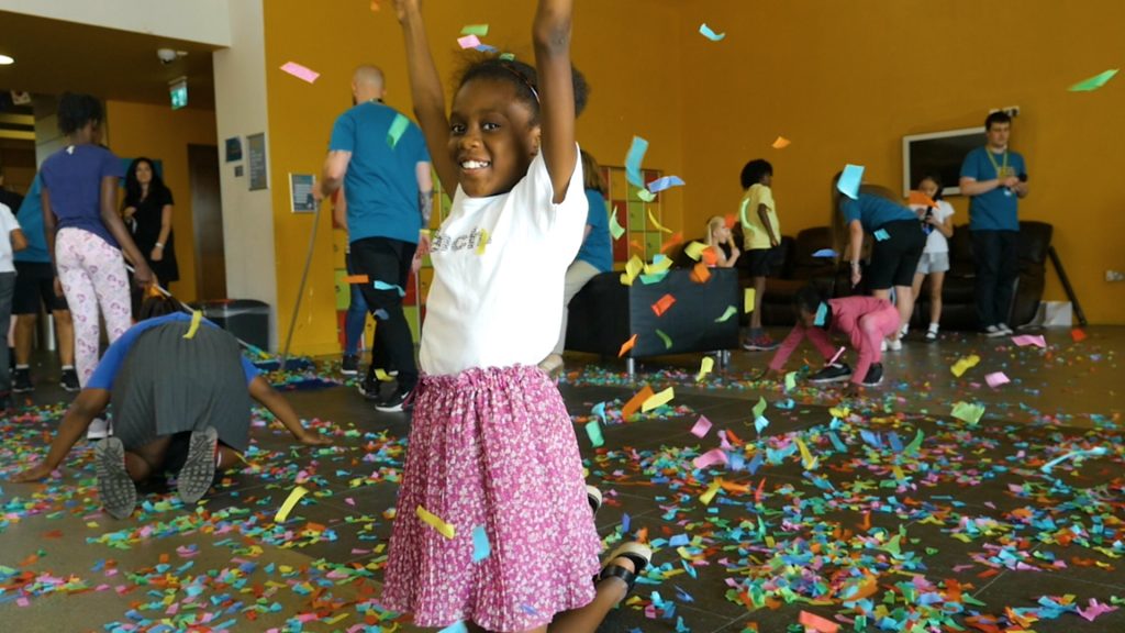 Young girl smiling with raised arms, confetti around her, and more children in the background - at Manchester Youth Zone