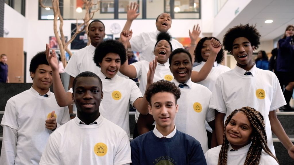 Photo of school children smiling, some with arms in the air, looking into the camera. Most are wearing white t-shirts with the We Rise logo