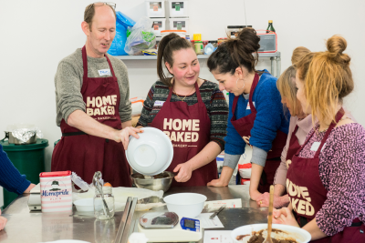 5 people wearing 'Home Baked' aprons gather round a work bench, working together to bake produce.