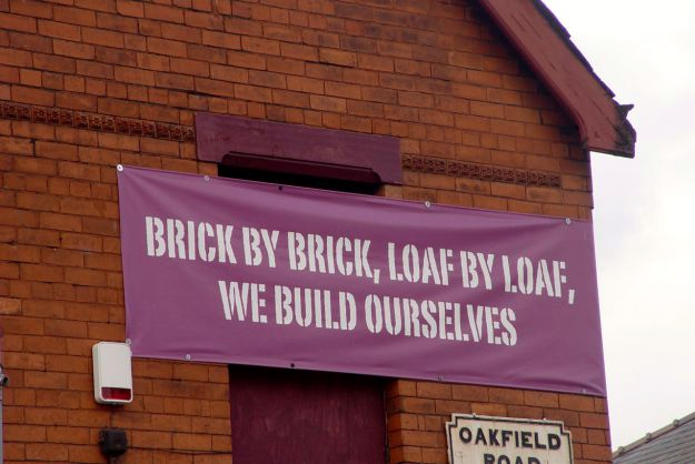 A Home Baked banner on a red brick building reads, "brick by brick, loaf by loaf, we build ourselves".