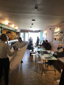 People in a the cafe and launderette. Some people are sitting at tables. Two people are folding a sheet together.